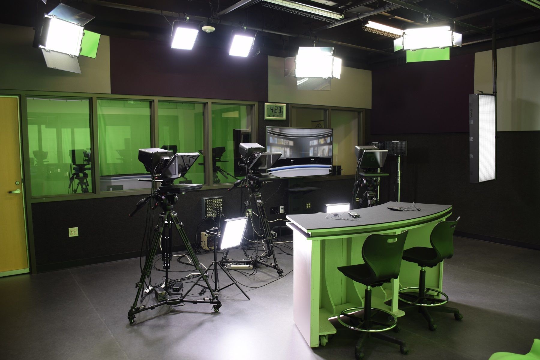 Empty TV studio with cameras, lighting, and green screens, featuring a presenter desk and chairs.
