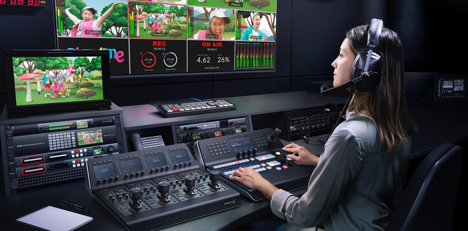 Person operating a control panel in a media production room with multiple screens displaying a colorful show.
