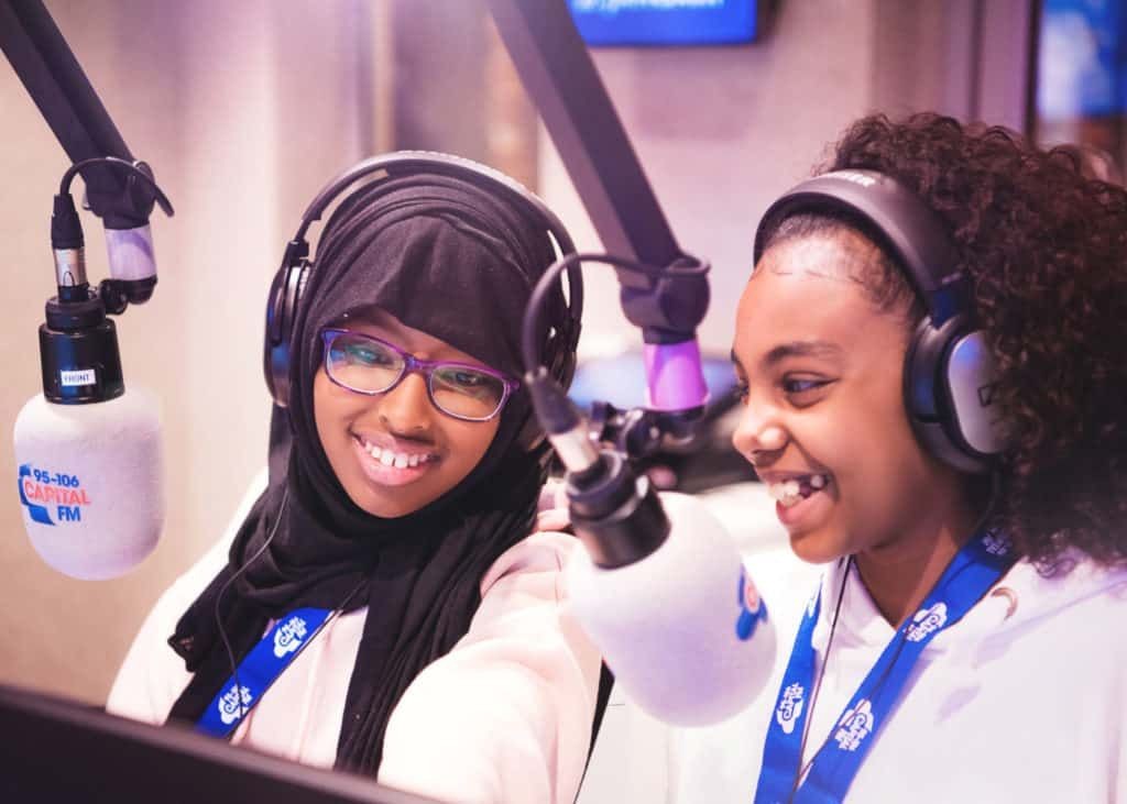 Two young girls smiling and speaking into Capital FM microphones in a radio studio.
