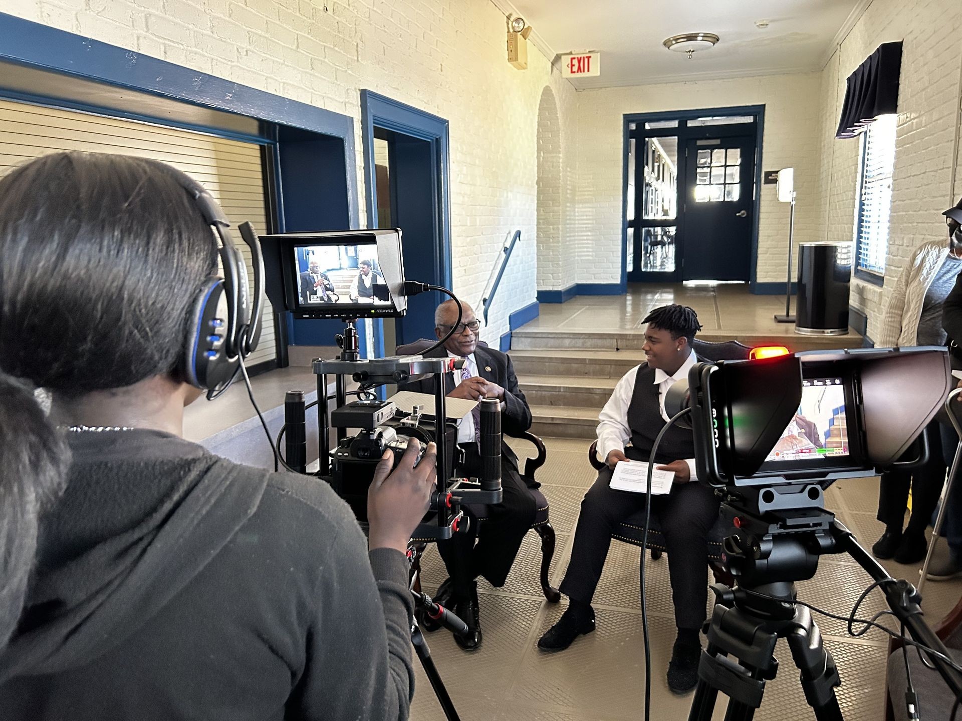 Camera crew filming two people sitting and talking in a hallway setting with audio and video equipment.